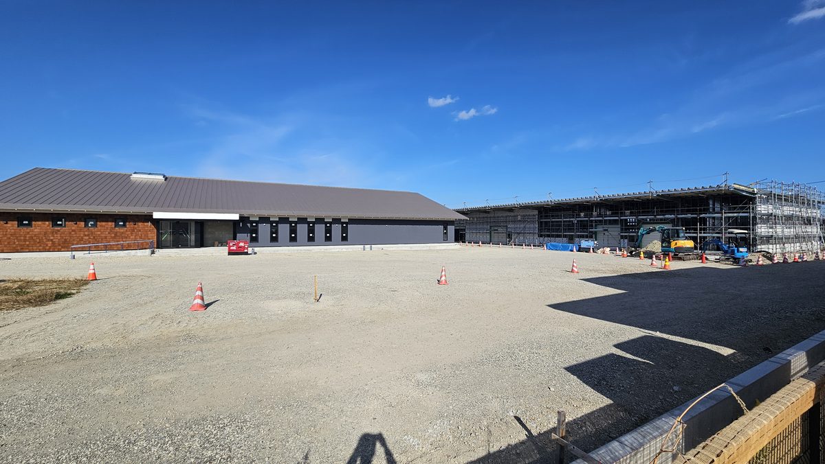 木目の外壁の建物の正面。ガラスの入口と手すり付きのスロープ、手前は砂利の広い敷地