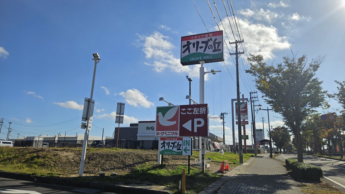 道路沿いに立つ「イタリア食堂 オリーブの丘 OLIVE HILL」の看板と、ヨークベニマルの「ここ左折 P」の看板の下にある「オリーブの丘 IN」の看板